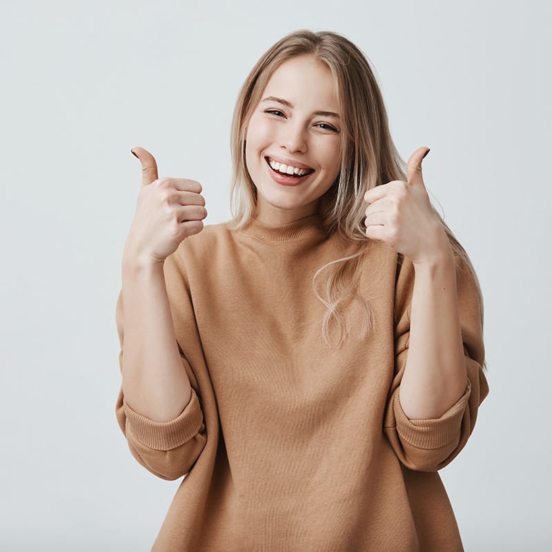 Portrait of fair-haired beautiful female student or customer with broad smile, looking at the camera with happy expression, showing thumbs-up with both hands, achieving study goals. Body language Rodzaje papierów i uszlachetnień ulotek w ofercie drukarni MadDruk – kreda mat, błysk i lakiery UV